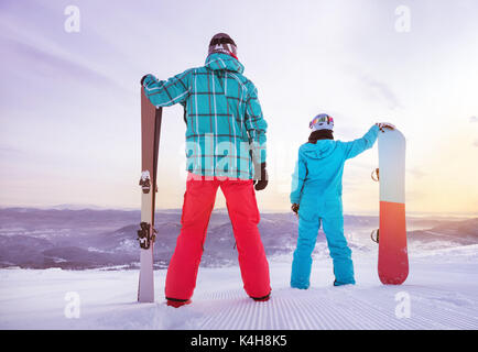 Familie von Vater und Tochter Skifahrer und Snowboarder steht auf Skipiste bei Sonnenaufgang oder Sonnenuntergang. Skifahren Konzept Stockfoto