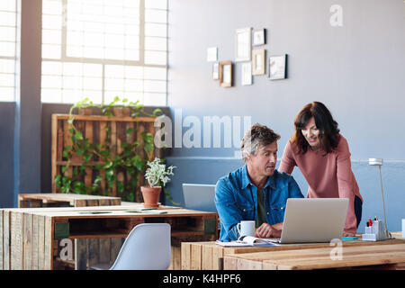 Zwei leger gekleidete junge Kollegen mit einem Laptop und miteinander sprechen, während Sie sich an einer Tabelle steht in einem modernen Büro Stockfoto