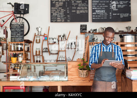 Lächelnden jungen afrikanischen Unternehmer Tragen einer Schürze gegen den Zähler seiner angesagten Cafes mit einem digitalen Tablet schiefen Stockfoto