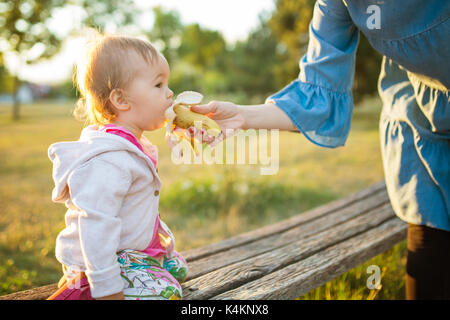 15 Monate altes Baby girl eathing die Banane aus der Hand ihrer Mutter; Hintergrundbeleuchtung, im Sonnenuntergang Stockfoto
