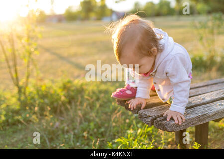 15 Monate altes Baby Mädchen klettern die Holzbank im Park Stockfoto