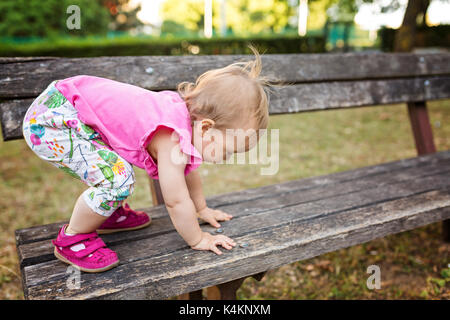 15 Monate altes Baby Mädchen klettern die Holzbank im Park Stockfoto