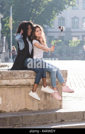 Lustige Freundinnen Spaß indem Sie Fotos mithilfe von selfie Stick beim Sitzen auf dem Brunnen in der Innenstadt. Stockfoto