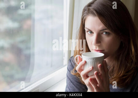 Schöne junge Frau sitzt am Fenster in heißes Getränk. Soft Focus Stockfoto