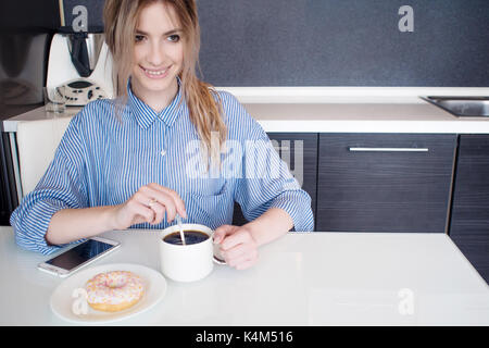 Lächelnde junge blonde Frühstück zu Hause essen. Kaffee mit Donut. Stockfoto