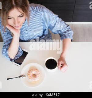 Charmante junge blonde Frühstück zu Hause essen. Kaffee mit Donut. Ansicht von oben Stockfoto