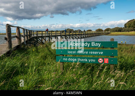 Menschen zu Fuß auf hölzernen Fußgängerbrücke über das Watt bei Flut, Aberlady Bay Nature Reserve, East Lothian, Schottland, Großbritannien, mit Schild, keine Hunde Stockfoto