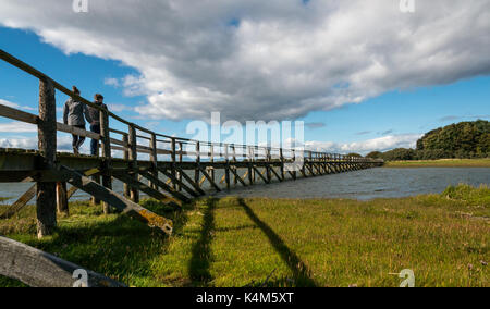 Junges Paar zu Fuß über Holzsteg über Wattenmeer bei Flut, Aberlady Bay Local Nature Reserve, East Lothian, Schottland, Großbritannien Stockfoto