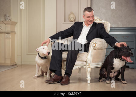 Der Mann Hundebesitzer petting zwei Hunde. Schwarz Pit Bull Terrier und Weiß oder staphorshire bulterrier sind im Vintage Interieur. Hunde sitzen auf beiden Seiten Stockfoto