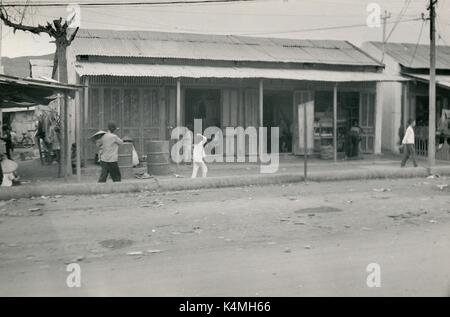 Street Scene mit Frauen und Kindern zu Fuß hinter einem Wellblech Gebäude auf einem staubigen Feldweg in Vietnam während des Vietnam Krieges, 1968. Stockfoto