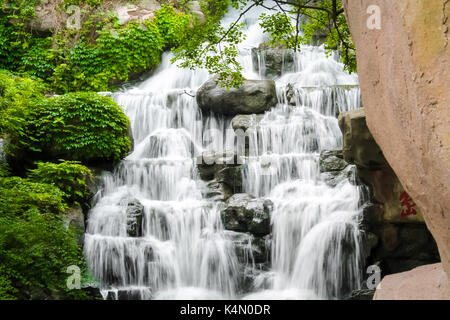 Wasser, das die Felsen in chinesischen Parks mit grünen Büschen und Stein im Hintergrund Stockfoto