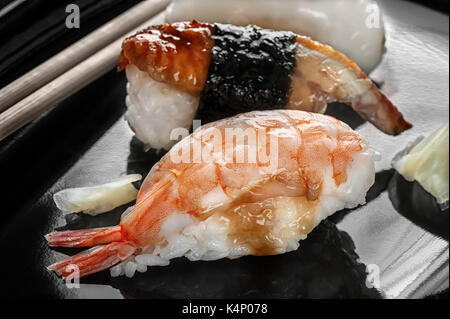 Japanisches Sushi aus Reis und Wolfsbarsch, Garnelen und geräuchertem Aal mit Stäbchen auf schwarzem Teller. Stockfoto