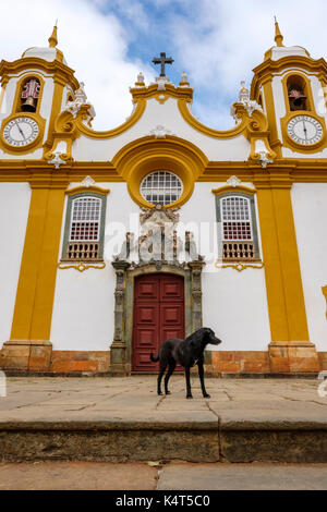 Schwarzer streunender Hund vor Matriz de Santo Antonio Kirche, der ältesten und größten katholischen Tempel von Tiradentes, Minas Gerais, Brasilien. Stockfoto