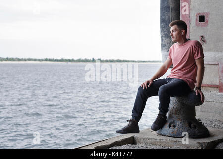 Der Typ hört Musik sitzen auf der Pier. Er sieht nachdenklich auf den Fluss. Jugend Mode. Stockfoto