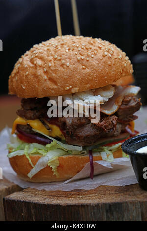 Große amerikanische Burger mit gezogenes Schweinefleisch, Zwiebelringe, Käse und Salat in Sesam Brötchen auf Pergament Papier über natürliche Holz geschnitten, Nahaufnahme, niedrigen Winkel Stockfoto