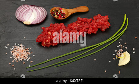 Rohes Hackfleisch rotes Fleisch Schnitzel, Gewürze, Pfeffer in Holz- Schaufel, spring green Schnittlauch, Zwiebeln, Knoblauchzehen und Himalaya Salz auf schwarzem Schiefer, Stockfoto