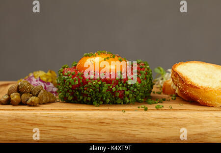 Eine Portion rohes Hackfleisch Fleisch Steak Tartar mit Eigelb, Baguette, grüne Schnittlauch, Zwiebeln und Kapern auf Holzbrett, Nahaufnahme, niedrigen Winkel v Stockfoto