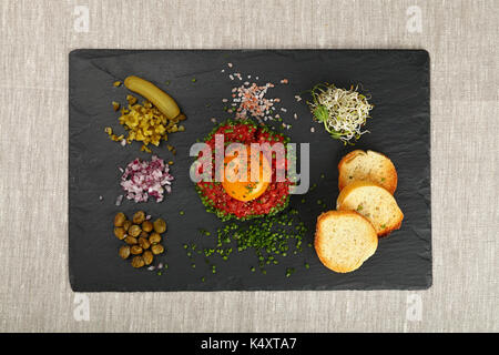 Eine Portion rohes Hackfleisch Fleisch Steak Tartar mit Eigelb, Grün, Schnittlauch, Zwiebeln, Gurken, Kapern, Salz und Toast am schwarzen Schiefer Board auf Tisch Stockfoto