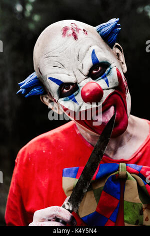 Nahaufnahme der Ein unheimlich bösen Clown tragen ein schmutziges rot Kostüm lecken ein großes Messer im Wald bei Nacht Stockfoto