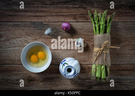 Bündel frischer Spargel mit Eiern, Zwiebeln und Salz auf rustikalen Holzmöbeln Hintergrund. Zutaten bereit, um eine Mahlzeit zu machen. Ansicht von oben. Stockfoto