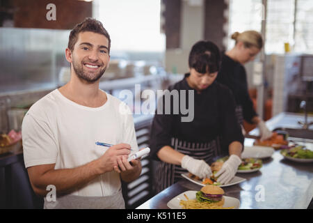 Portrait von lächelnden Kellner stehen gegen weibliche Chefs die Zubereitung von Speisen in der Küche im Café Stockfoto