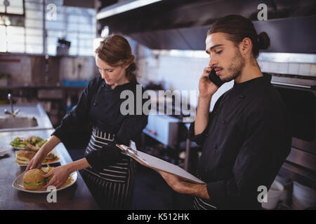 Junger Kellner sprechen auf dem Smartphone während Kellnerin die Zubereitung von Speisen in der gewerblichen Küche im Café Stockfoto