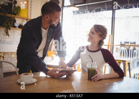 Junger Mann mit Tablet-PC im Stehen durch Frau am Tisch sitzen im Café Stockfoto