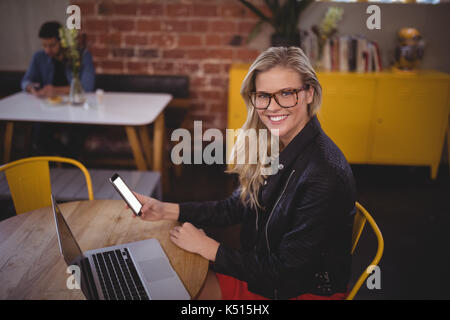 Porträt der lächelnde junge blonde Frau mit Smartphone beim Sitzen mit Laptop am Tisch im Café Stockfoto