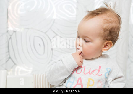 Elf Monate alten Baby Mädchen in einem Hocker, lustige Gesichter in die Kamera sitzt, überrascht und ihr Finger klemmt im Mund Stockfoto