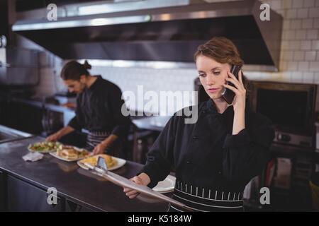 Junge Kellnerin reden auf dem Smartphone während der Kellner die Zubereitung von Speisen in der gewerblichen Küche im Café Stockfoto