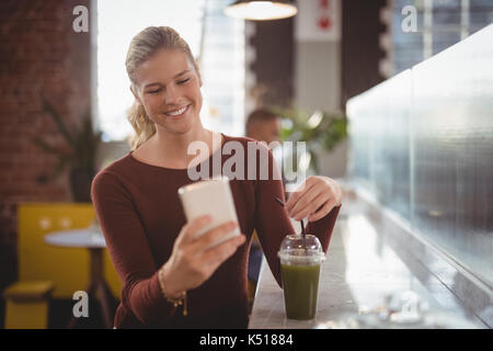 Lächelnde junge blonde Frau mit Smartphone während der Sitzung mit Getränk an der Theke im Café Stockfoto