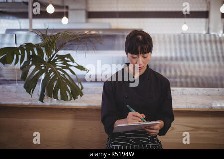 Junge Kellnerin schreiben auf Zwischenablage während gegen Counter im Café sitzen Stockfoto