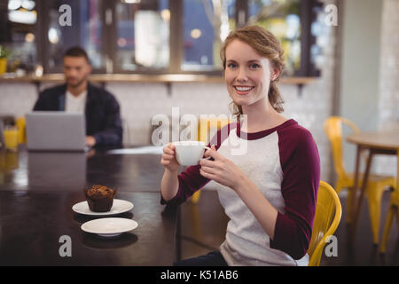 Portrait von lächelnden jungen Frau mit Tasse Kaffee beim Sitzen am Tisch im Cafe Stockfoto