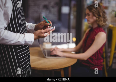 Mittelteil der Kellner schreiben auf Notepad gegen weibliche Kunden am Tisch sitzen im Café Stockfoto