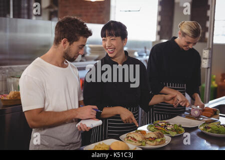 Fröhlicher Koch an Nahrungsmittel, die von den weiblichen Kollegen im Café Küche, Kellner Stockfoto