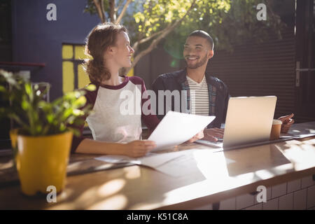 Lächelnde junge kreative Fachleute sprechen beim Sitzen mit Laptop am Tisch im Café Stockfoto
