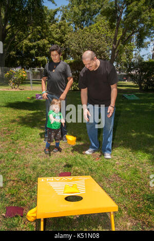 Mit seinem Vater und seinem Bruder aufpassen, ein Spanischer Junge wirft eine Bohne Beutel an einem gelben Ziel während einer Costa Mesa, CA, Festival in einem Stadtpark. Stockfoto