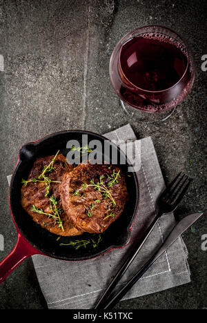 Für eine Person ein Mittagessen. Hausgemachte gegrillte Rindersteaks mit Thymian in einer Portionierten Pfanne, mit einer Gabel, Messer und einem Glas Wein. Auf schwarzen Stein Tabelle Stockfoto