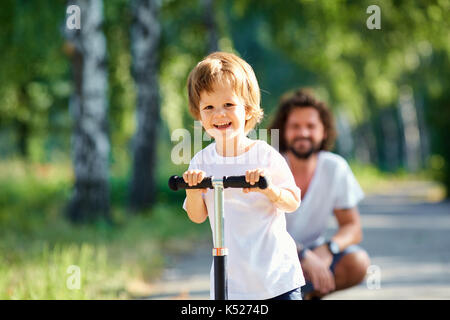 Der Junge Rochen auf einem Roller im Park mit seinem Vater Stockfoto