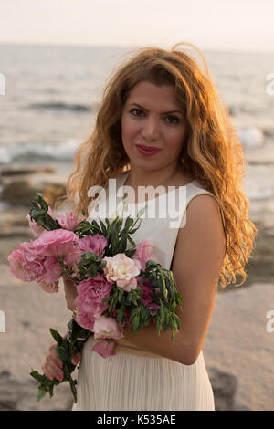 Schöne Frau mit lockigem Haar trägt weißes Kleid und hält Bouquet von rosa lisianthus Stockfoto