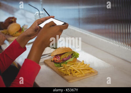 7/8 Hände von Frau fotografieren Pommes und Burger an der Theke im Cafe Stockfoto