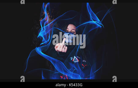 Rebellische Jugend: Mädchen mit einem Schädel Maske zeigt die mittleren Finger auf einen dunklen Hintergrund mit blauen Flammen des Lichtes Maltechnik. Stockfoto
