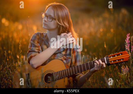 Schönen kaukasischen junge Frau in die Gläser und Plaid Shirt mit einer akustischen Gitarre auf einer Wiese im Abendlicht. Herbst Spaziergang im Freien Stockfoto