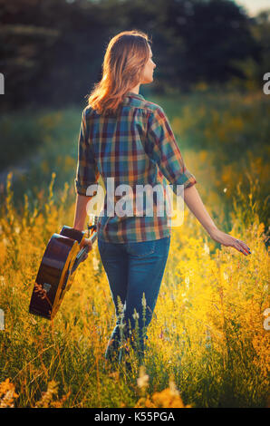 Junges Mädchen mit einer akustischen Gitarre Spaziergänge durch den Herbst im Freien bei Sonnenuntergang. Ansicht von der Rückseite. Stockfoto