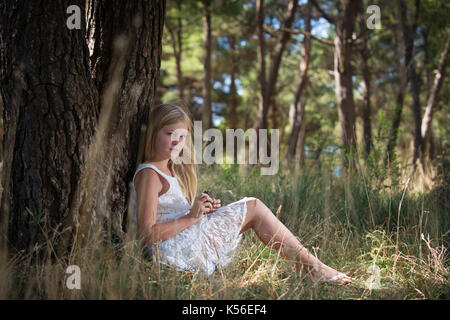 Lange Blonde süße Mädchen in weißem Kleid, lehnte sich mit dem Rücken gegen den Baum mit Pine Cone in Händen. Märchen sehen gleich aus. Stockfoto