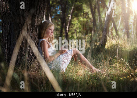 Lange Blonde süße Mädchen in weißem Kleid, lehnte sich mit dem Rücken gegen den Baum mit Pine Cone in Händen. Märchen sehen gleich aus. Stockfoto