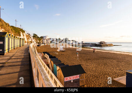 Am frühen Morgen, lange Schatten, Blick entlang Strandhütten und Strand in Viking Bay Broadstairs mit Hafen im Hintergrund. Strahlender Sonnenschein. Stockfoto