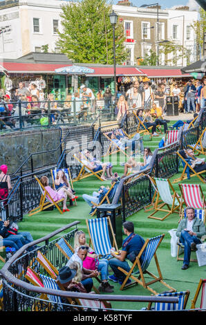 Die Leute sitzen und in Liegestühlen im Camden Market entspannen, Die Stühle sind auf Kunstrasen auf verschiedenen Ebenen eines Farbverlaufs einstellen. Stockfoto