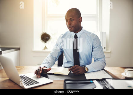 Junge afrikanische Unternehmer trägt ein T-Shirt und an seinem Schreibtisch in einem Büro online arbeiten mit einem Laptop Riegel ausgerichtet Stockfoto