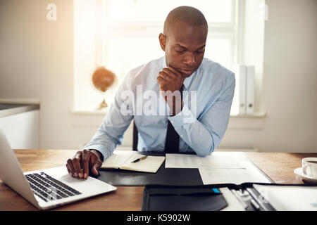 Junge afrikanische Unternehmer Lesen von Dokumenten und arbeitet an einem Laptop, während an seinem Schreibtisch in einem Büro sitzen Stockfoto
